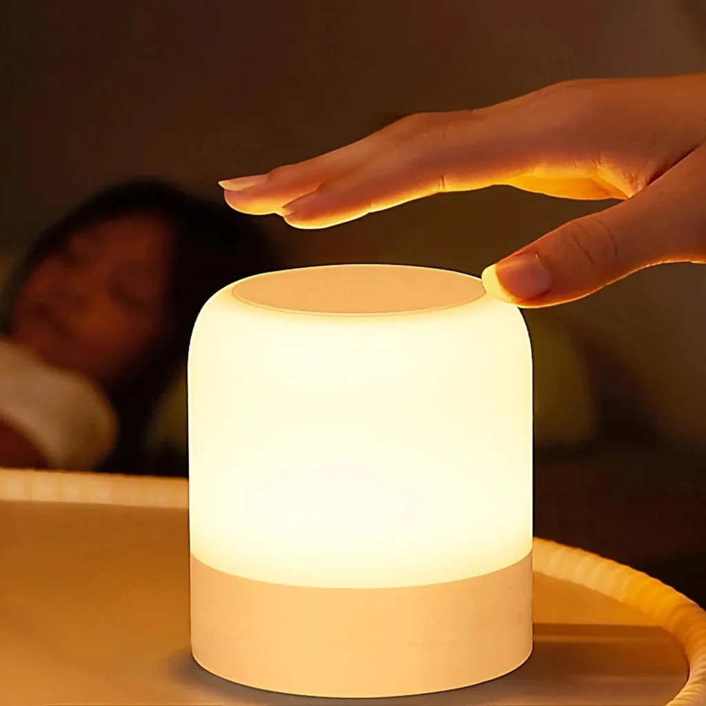 Close-up of a hand tapping the top of a cylindrical night light with a wooden base to adjust brightness, with a sleeping child in the background.